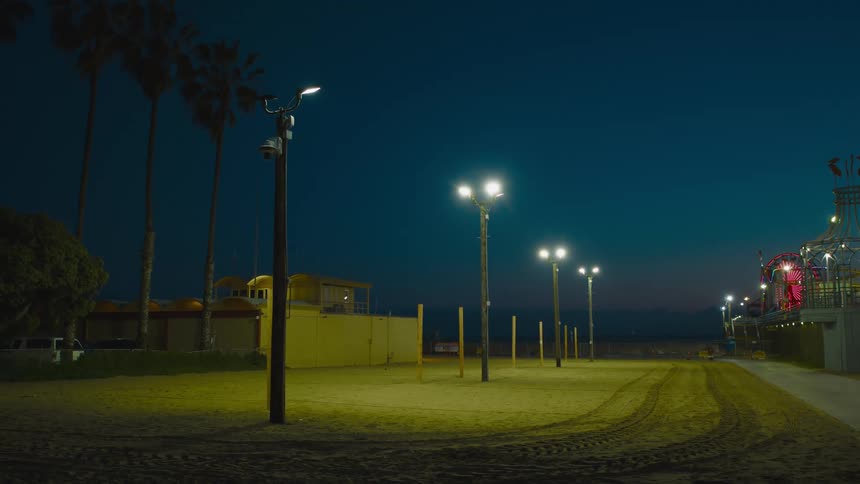 Lights reveal vehicle tracks along dirt between park building and nearby amusement park with lit ferris wheel under night sky