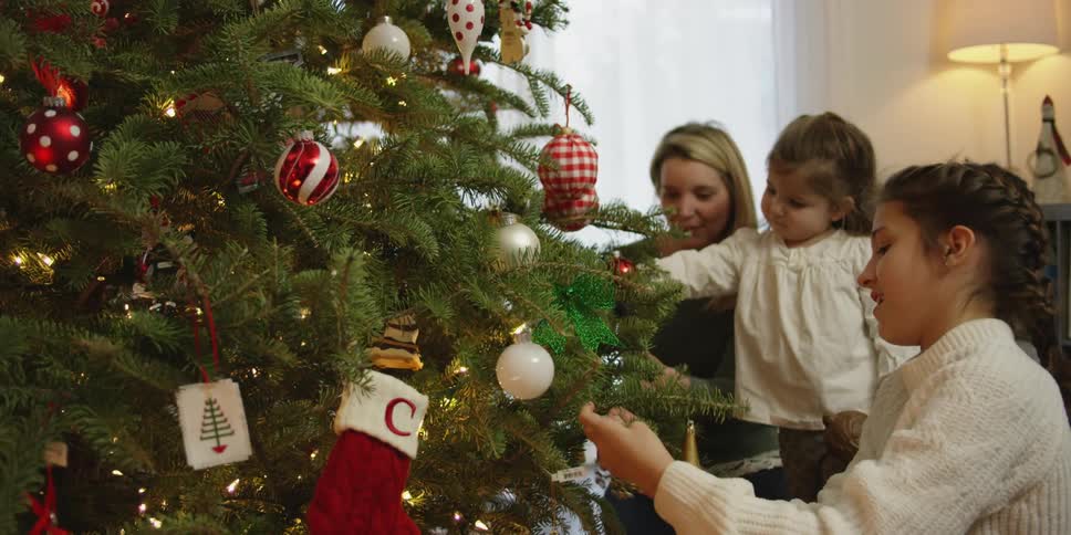 A mom and her two daughter trim the christmas tree at home with sun shining through the sheers