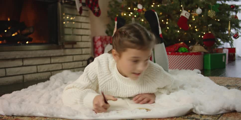 A girl in a room laying on a blanket writing on a book with a christmas tree and a fireplace in the background