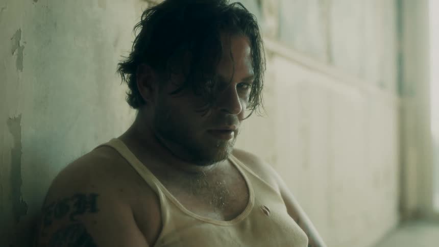 A man in a dingy tank top sits against the wall in a bare dingy room