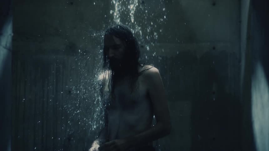 A man reaches up to touch his long hair as water falls down in a concrete walled shower