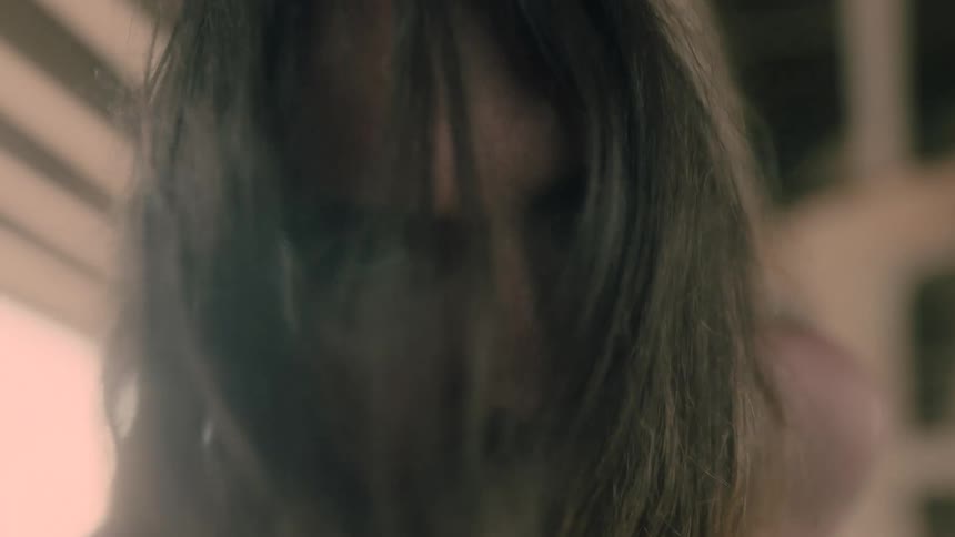 A man with long hair and a beard twitches while staring aggressively at the camera