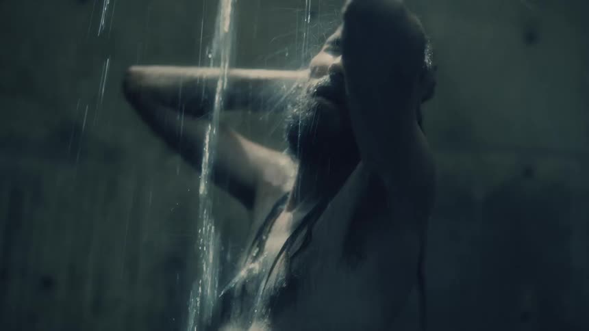A man with long hair and beard shakes his head and sways as water showers down from above