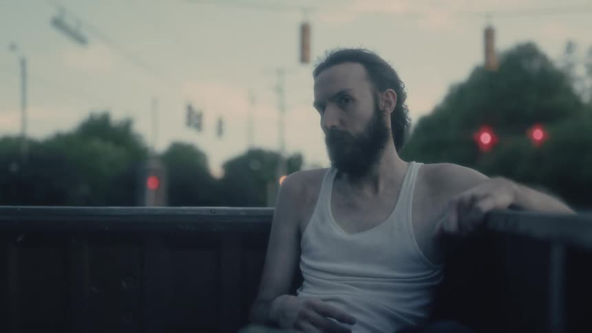 A thin man with a long beard rides in the back of a pickup truck