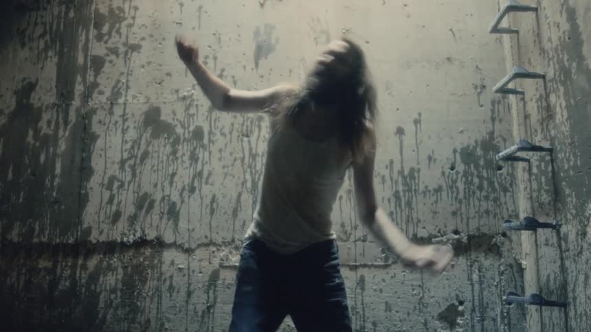 A lean and scruffy man yells in a fit of anger while he stands a room with concrete walls