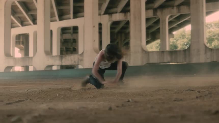 A man with long hair and a beard thrashes around on the dirt beneath a concrete overpass