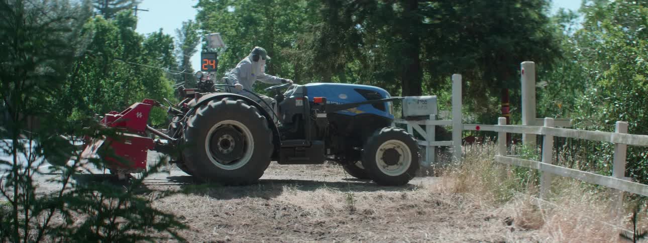 Cars drive down a rural street as a man climbs on a tractor and drives it in to a field