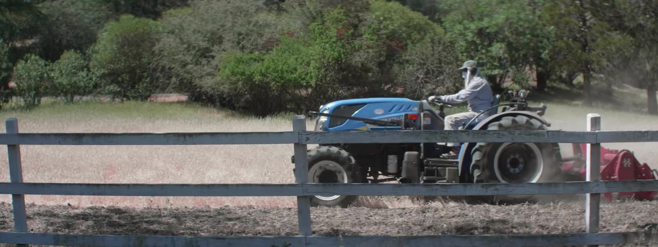 A person driving a tractor with a fence in the foreground and with trees in the background