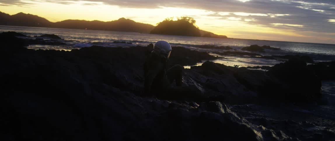 Waves crash against rocks on the shore and a person with a helmet on gets up and runs