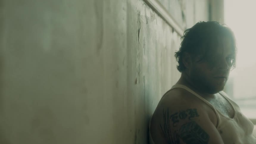 An overweight man in a tank top sits against a wall and looks upset