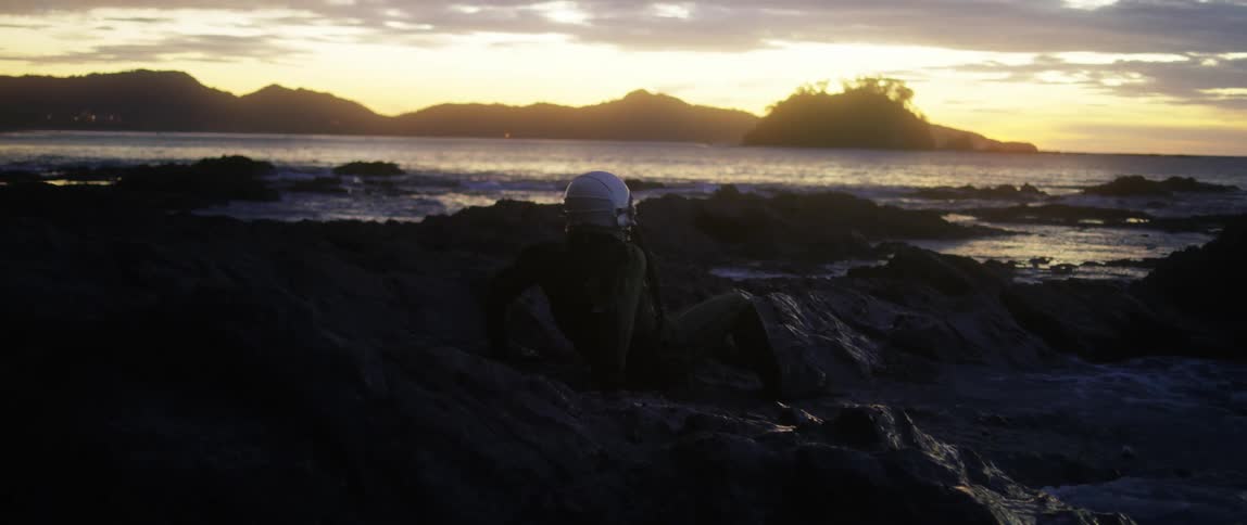 Waves crash against rocks as a person with a helmet lying in the water gets up and starts to run