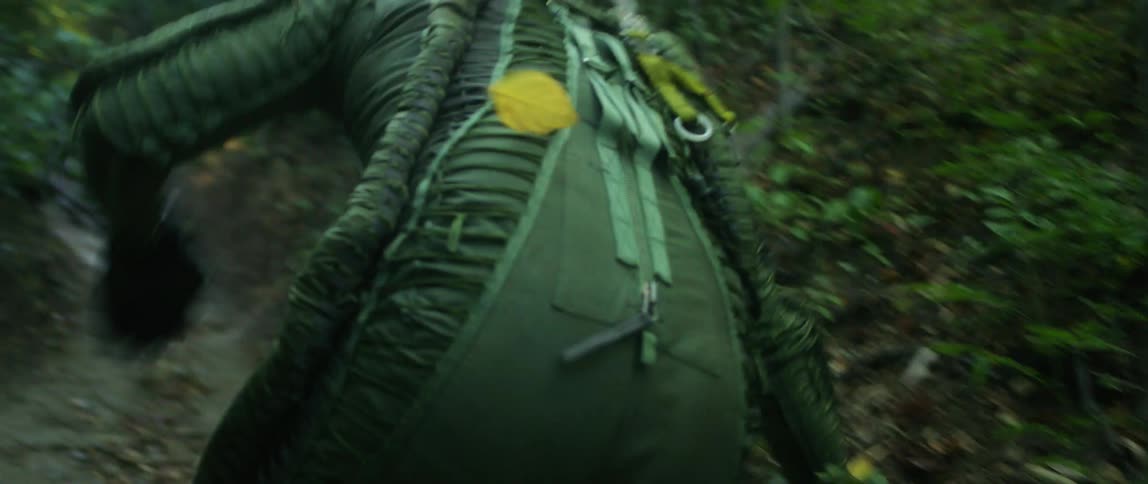 Man wearing pilot suit with white helmet slips as he runs through woods then regains his balance and keeps running