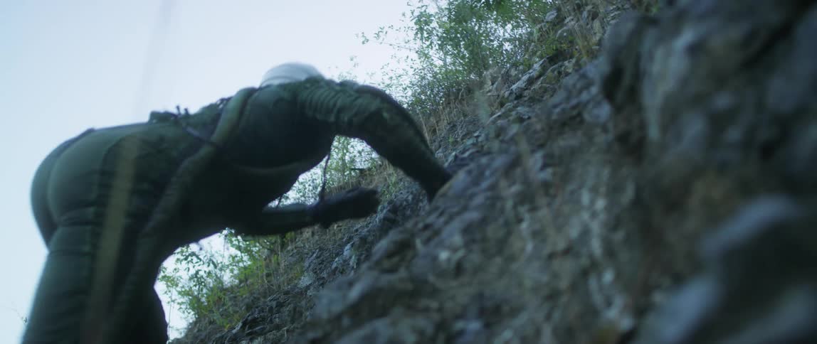 A hiker climbs a steep cliff on a mountainside at day and the hiker pushes down some rocks during the climb