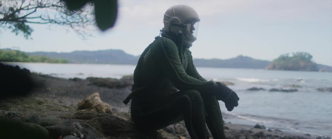 A man in a diving helmet and body suit sits on shore watching the waves