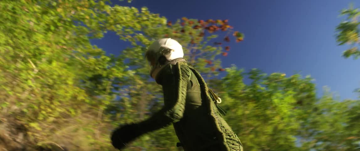 A man with a helmet and a flight suit walking in a pathway in the forest with the sky and clouds in the background