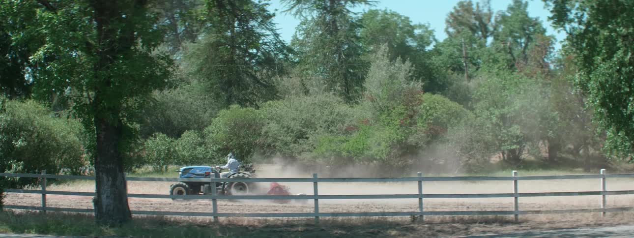 A person driving on a tractor with a fence and a road in the foreground and with trees the sky and clouds in the background