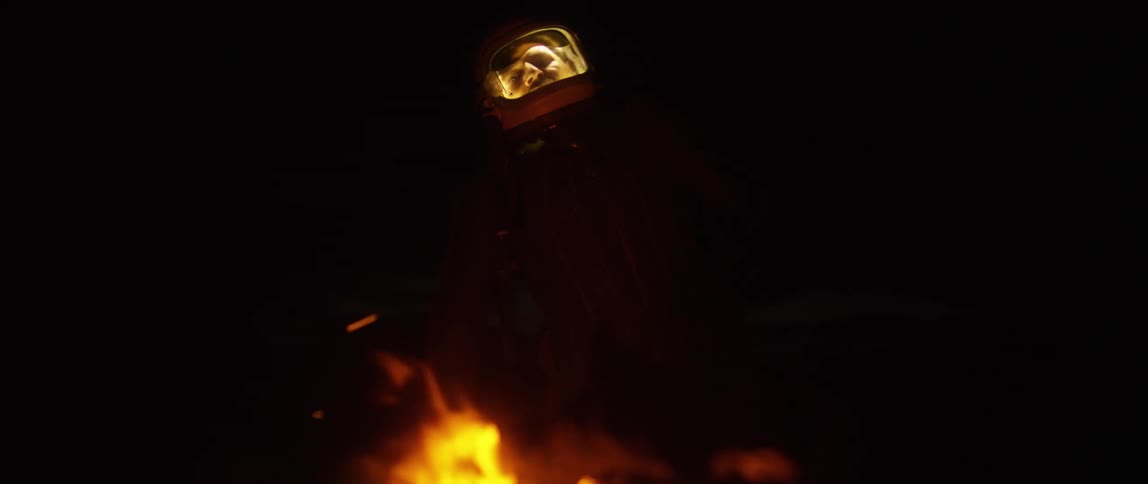 A person with a helmet and a flight suit with a bonfire burning in the foreground