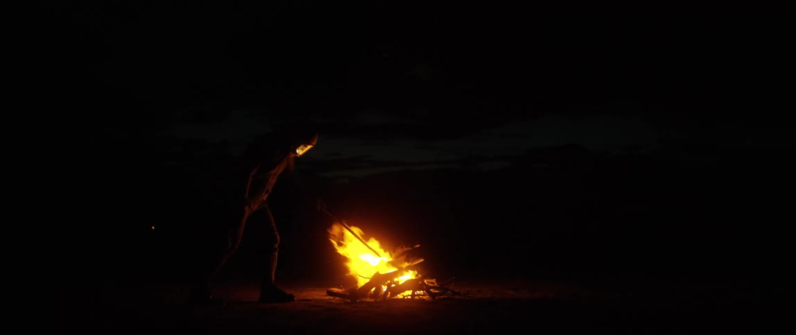 A figure in a helmet with visor pokes a burning camp fire at night