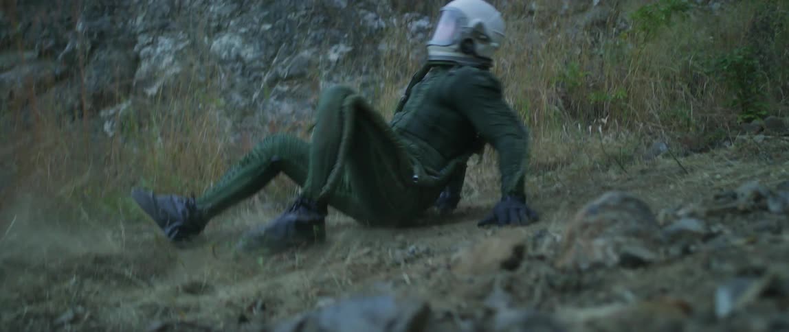 Person wearing white helmet and green jumpsuit slips and falls on ground at base of stone slope