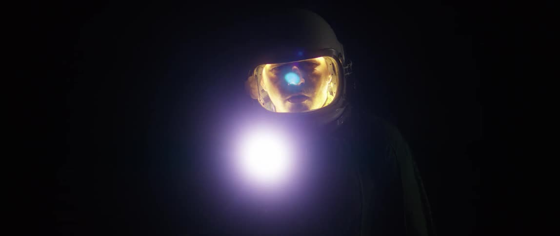 A man wearing an astronaut helmet which is illuminating his face walks carefully through a dark environment