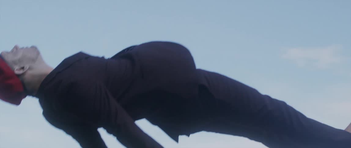 Barefoot man wearing suit and beret falls backwards under lightly clouded sky