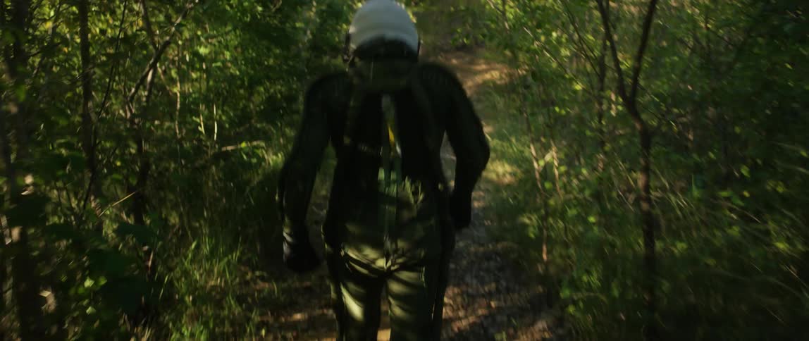 Man wearing green jumpsuit and white helmet walks along dirt path through woods