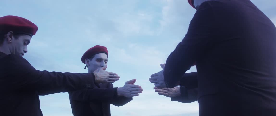 Four pale men in identical red hats stand in a circle making hand motions