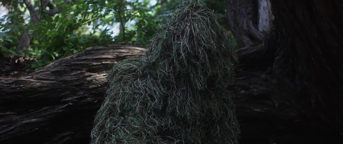 A person in a ghillie suit turns their head alertly while crouched in a forest
