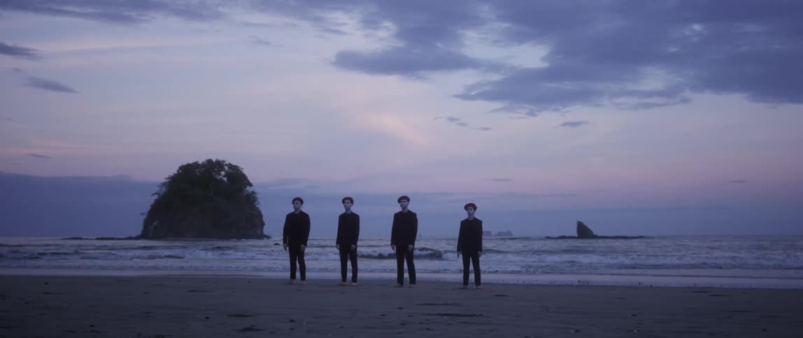 Four men in suits and berets stand in the sand facing away from the waves at dusk