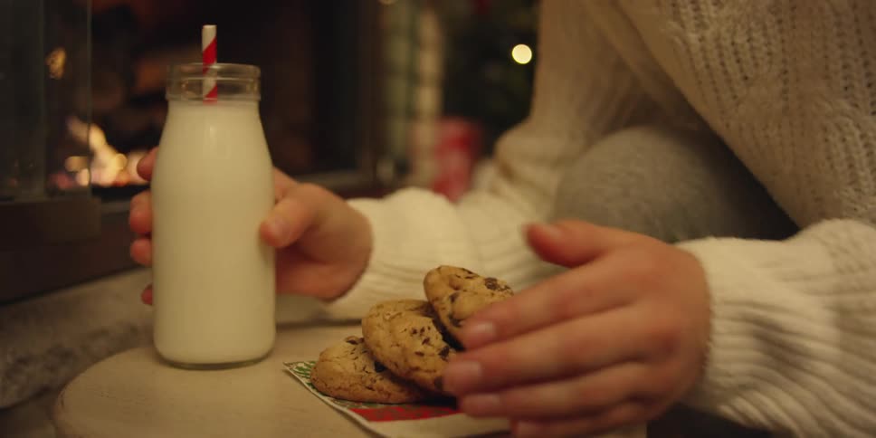 A person putting milk and cookies on a stool with a christmas tree and a fireplace in the background