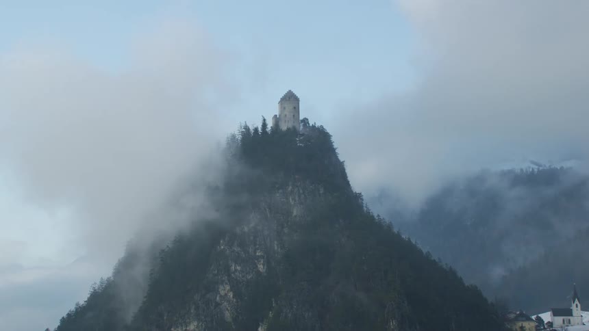 Tower sits on summit covered by snow and conifer forest as clouds pass while village sits near foot of mountain
