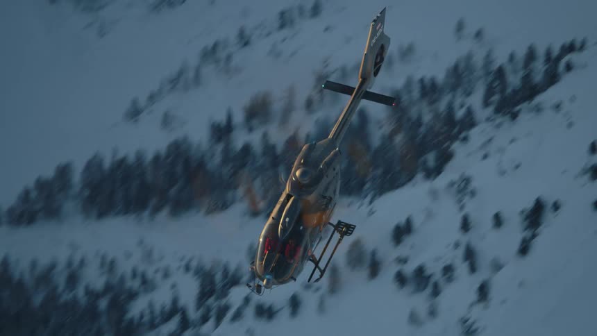 A helicopter flies over a snow covered slope dotted with trees