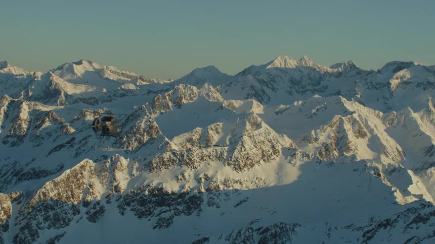 A helicopter flies over jagged rocky snow covered ridges and peaks