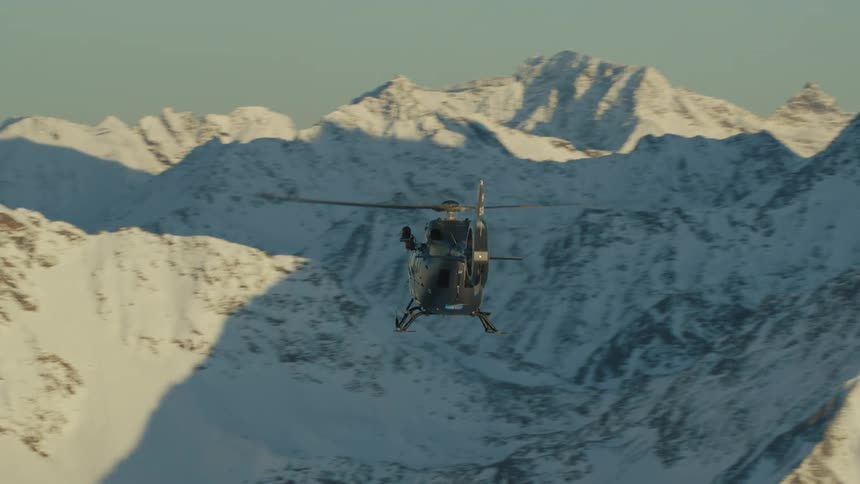 A helicopter flies and hover near snow covered jagged peaks