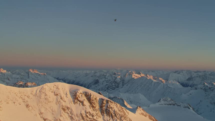 A helicopter flies above snow covered rocky ridges at dawn