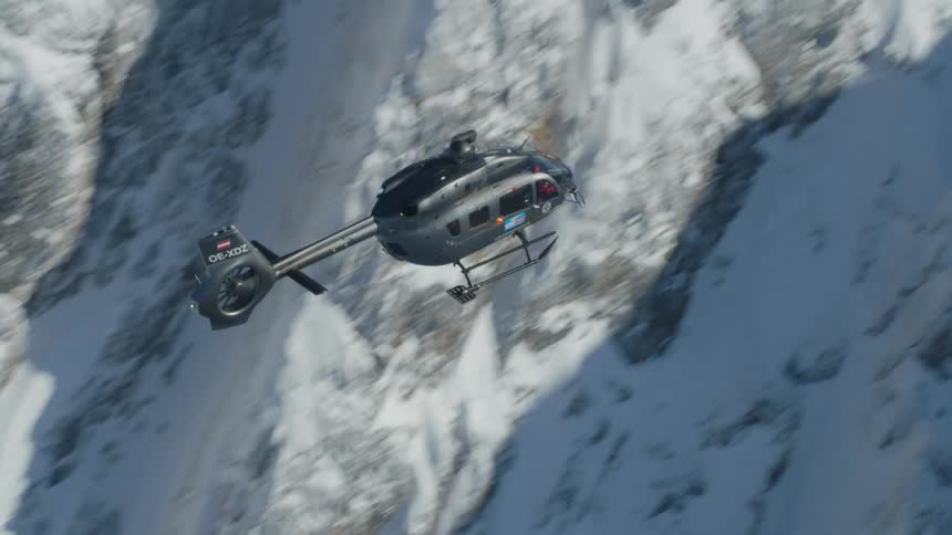 A helicopter flies over snow covered slopes and rocky ridges