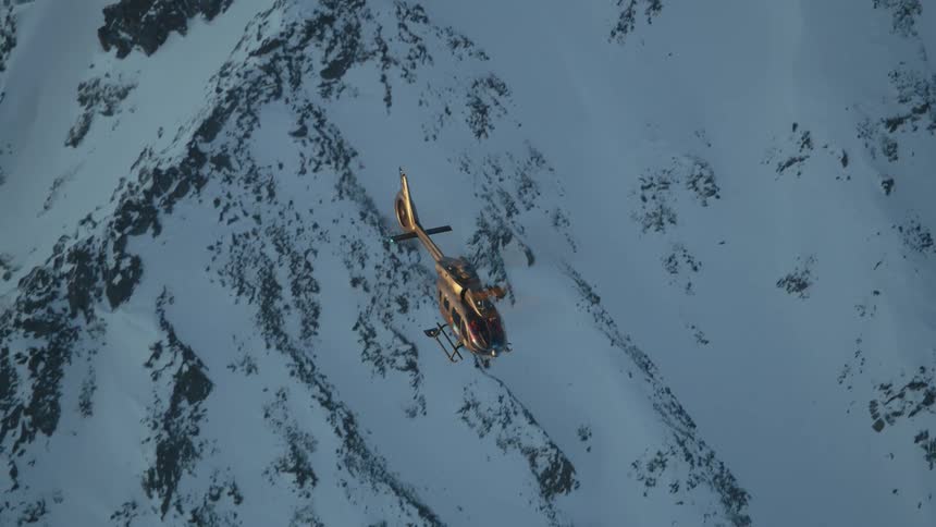 A dark helicopter flies over snow covered mountains
