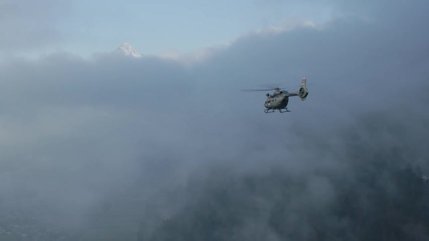 A helicopter travels through the fog obscuring a snow capped mountain range during the day