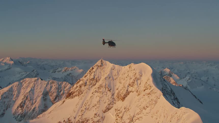 A helicopter flying over jagged snow covered ridges at dawn