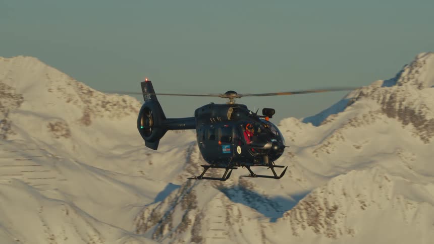 Pilot wearing red flies helicopter over snow covered mountains slopes under clear sky