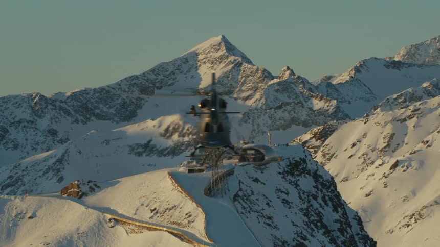 A helicopter flies above rocky snow covered ridges