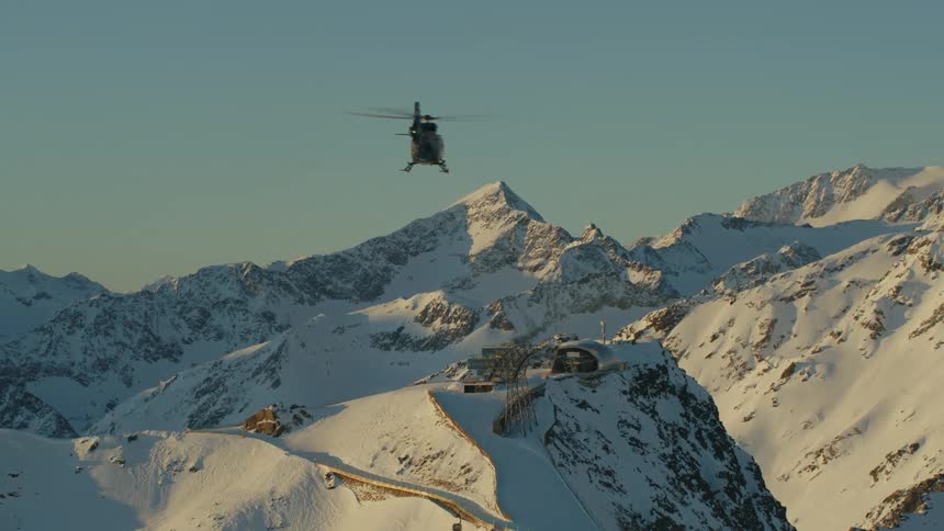 A helicopter flies over a ski resort on the top of snow capped mountains