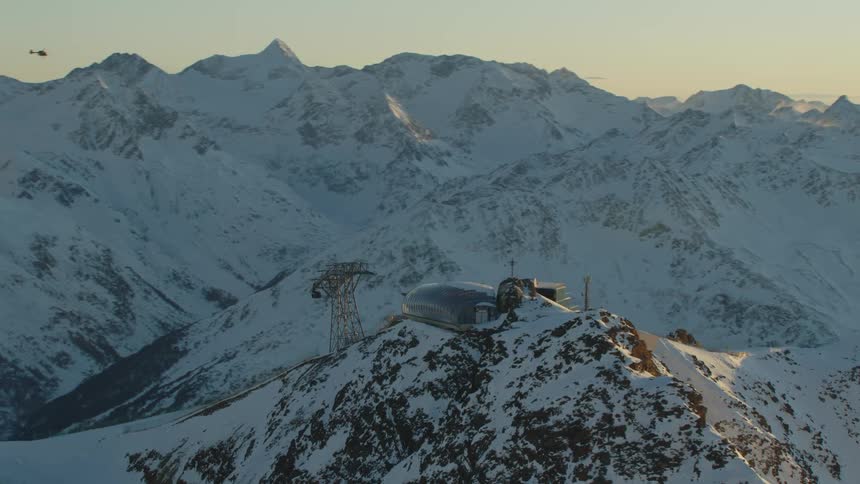 Helicopter hovers over mountains in background as cable car arrives at tower on snow covered mountain summit
