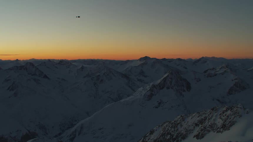 The silhouette of a helicopter flying above snow covered jagged ridges at sunrise