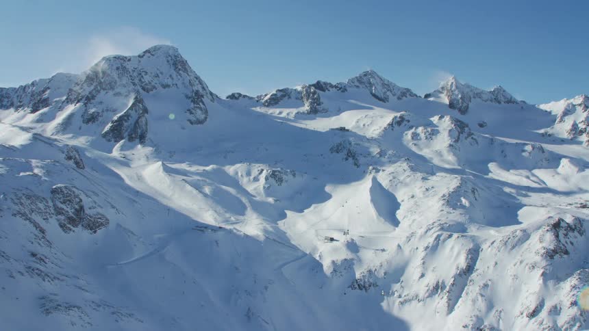 Wispy clouds hover over peaks in mountain range covered in snow and partially covered in shadow