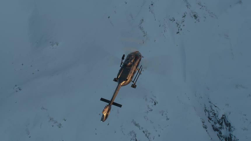 A helicopter glowing gold in the sunlight flies above snow covered slope