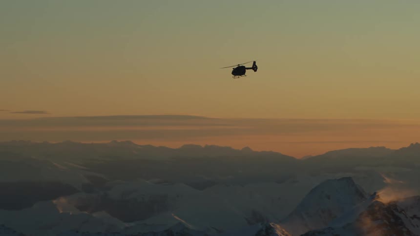 A helicopter flying over snowy mountains with an orange sky at sunset