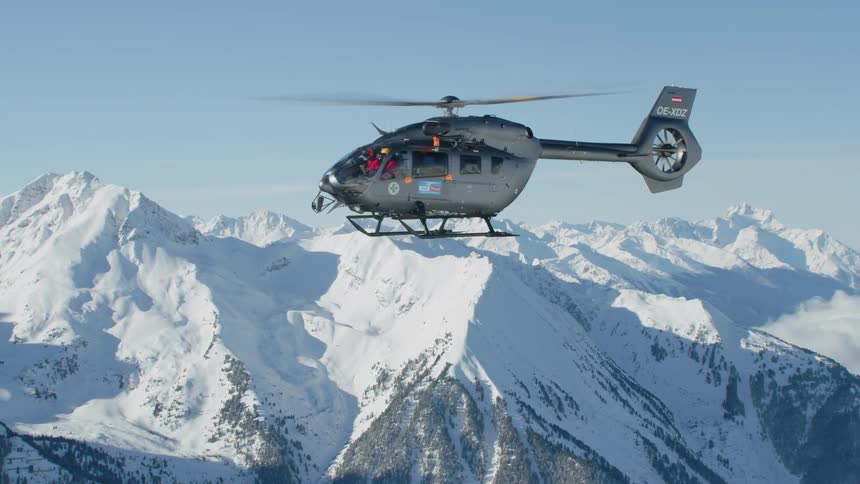 Pilot wearing red flies helicopter over snow and forest covered mountain range under a clear blue sky