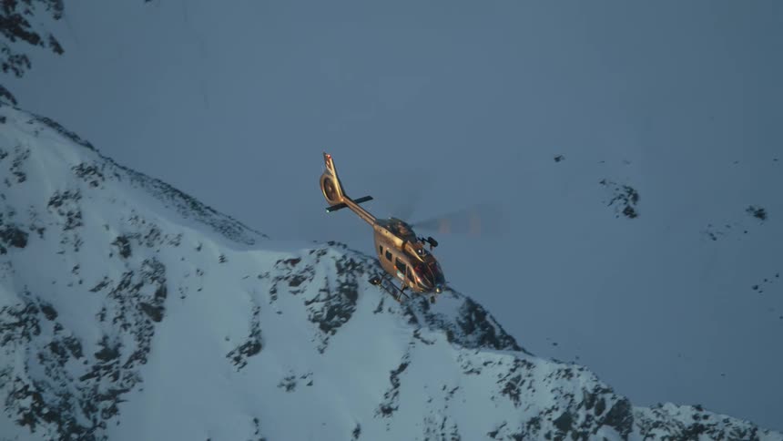 A helicopter flies sideways over snow covered mountains