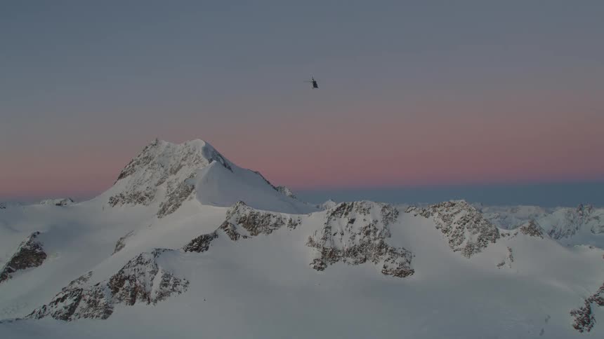 A helicopter flies over snow covered rocky ridge at dawn
